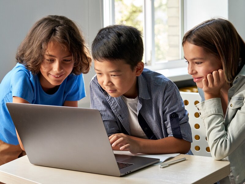 Drei Kinder sitzen gemeinsam an einem Tisch und schauen interessiert auf den Bildschirm eines Laptops.