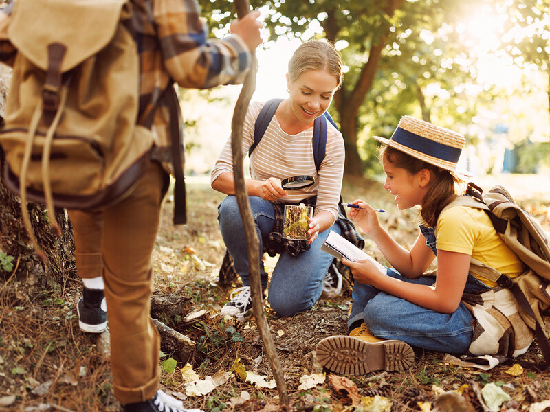 Eine Mutter und ihr Kind hocken auf dem Waldboden und betrachten Laub in einem Glas. Das Kind macht Notizen in einem Heft.