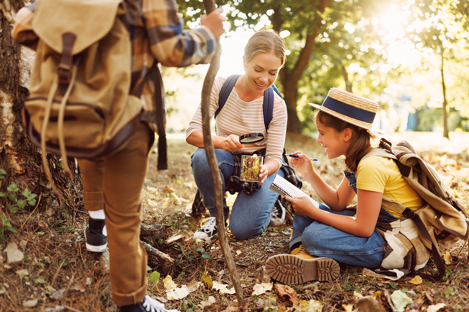 Eine Mutter und ihr Kind hocken auf dem Waldboden und betrachten Laub in einem Glas. Das Kind macht Notizen in einem Heft.