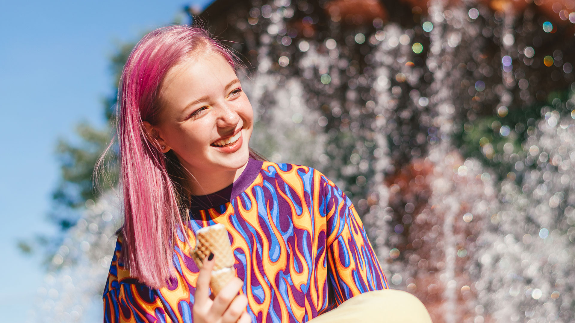Mädchen mit pink gefärbtem Haar lächelt, hinter ihr ein Springbrunnen und blauer Himmel