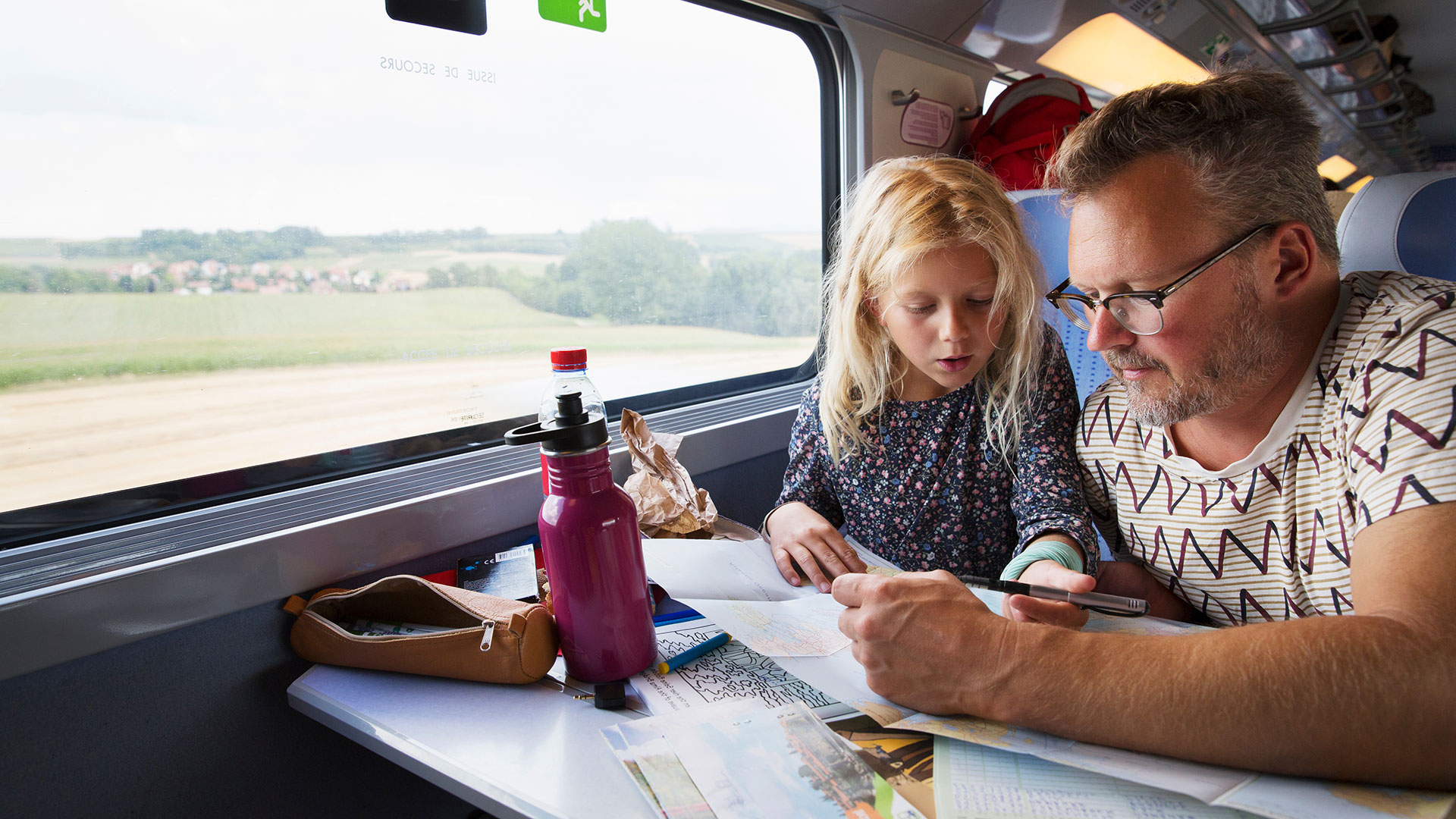 Mann mit Bart und ein Mädchen mit blonden Haaren sitzen an einem Tisch im Zug