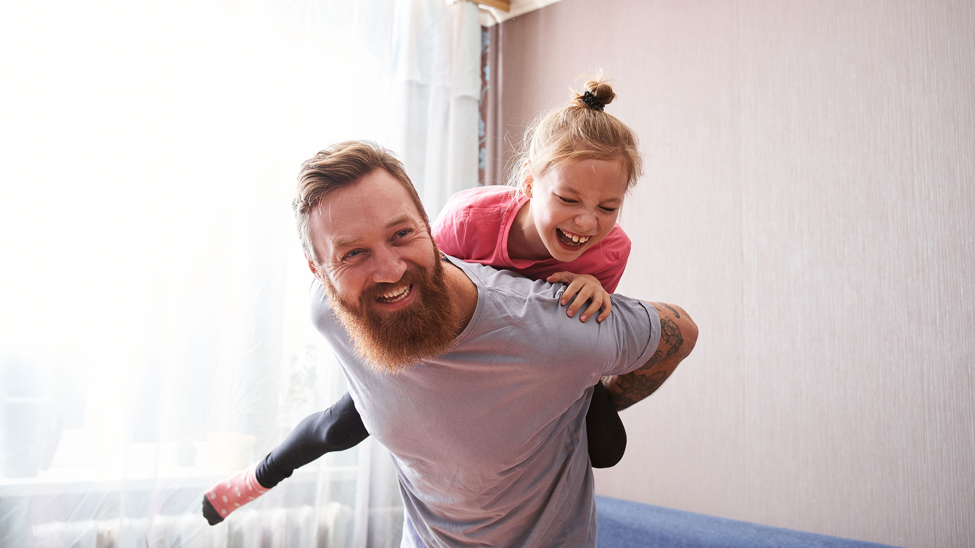 Mann mit Bart in einem Raum mit Couch, er trägt ein junges Mädchen auf dem Rücken, beide lachen