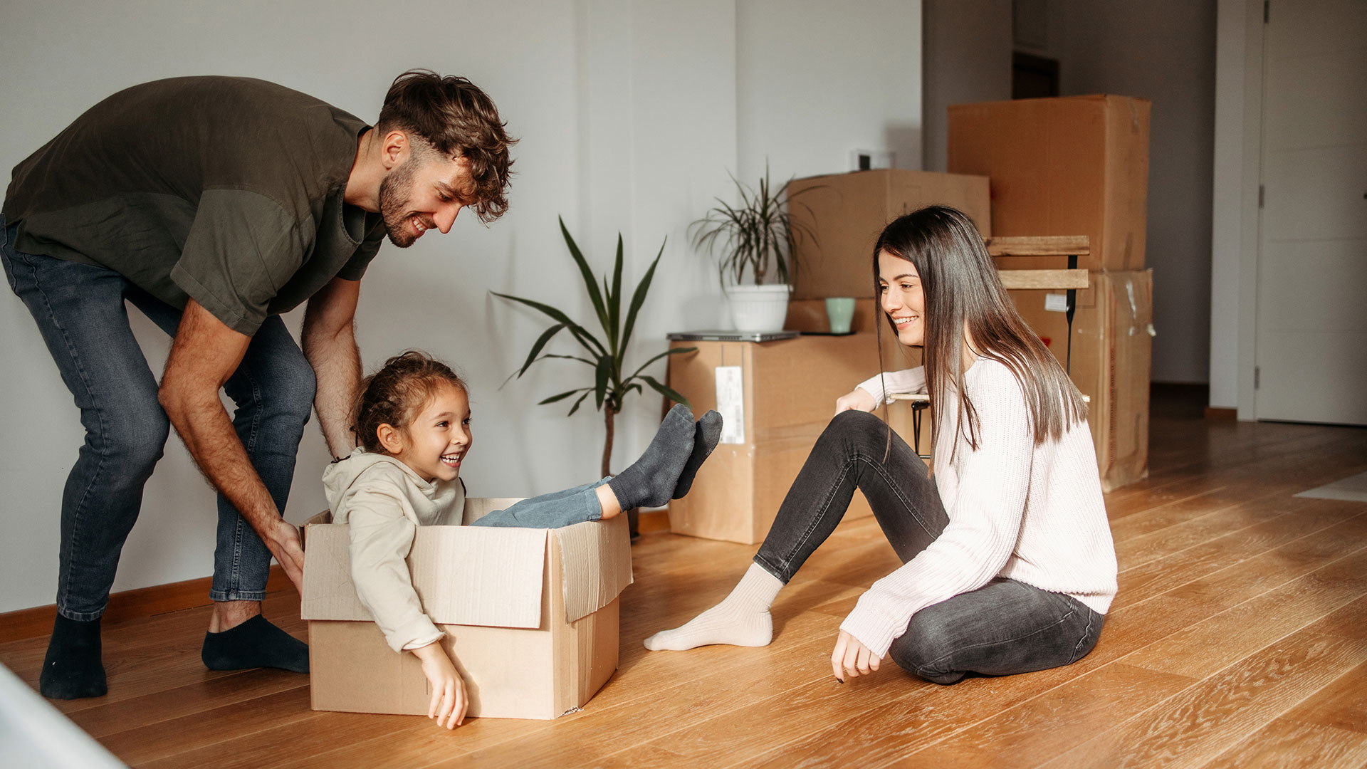 Ein junger Mann schiebt ein Kind, dass in einem Umzugskarton sitzt, durch eine leeren Wohnraum. Eine Frau sitzt daneben auf dem Parkettboden und schaut freudig zu