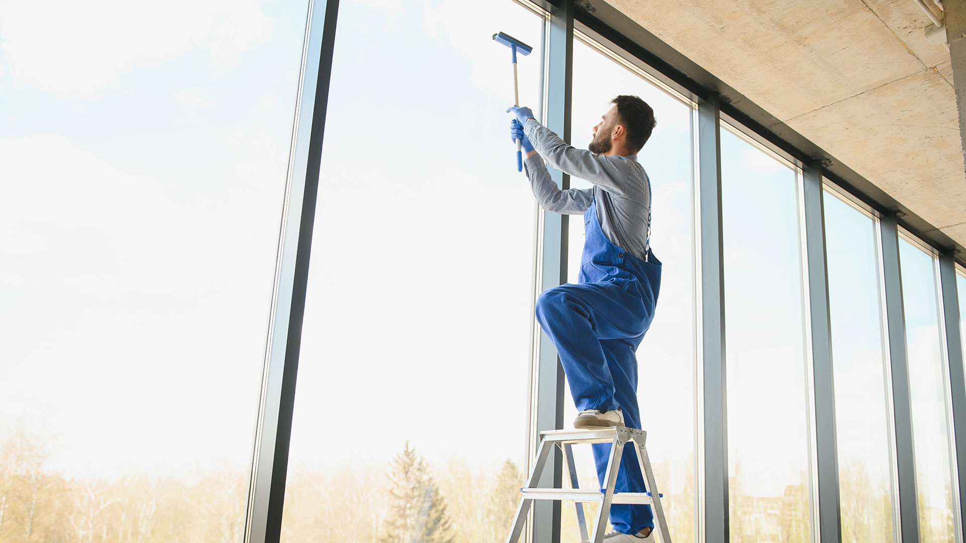 Mann an einer großen Fensterfront, steht auf einer Leiter und putzt ein Fenster