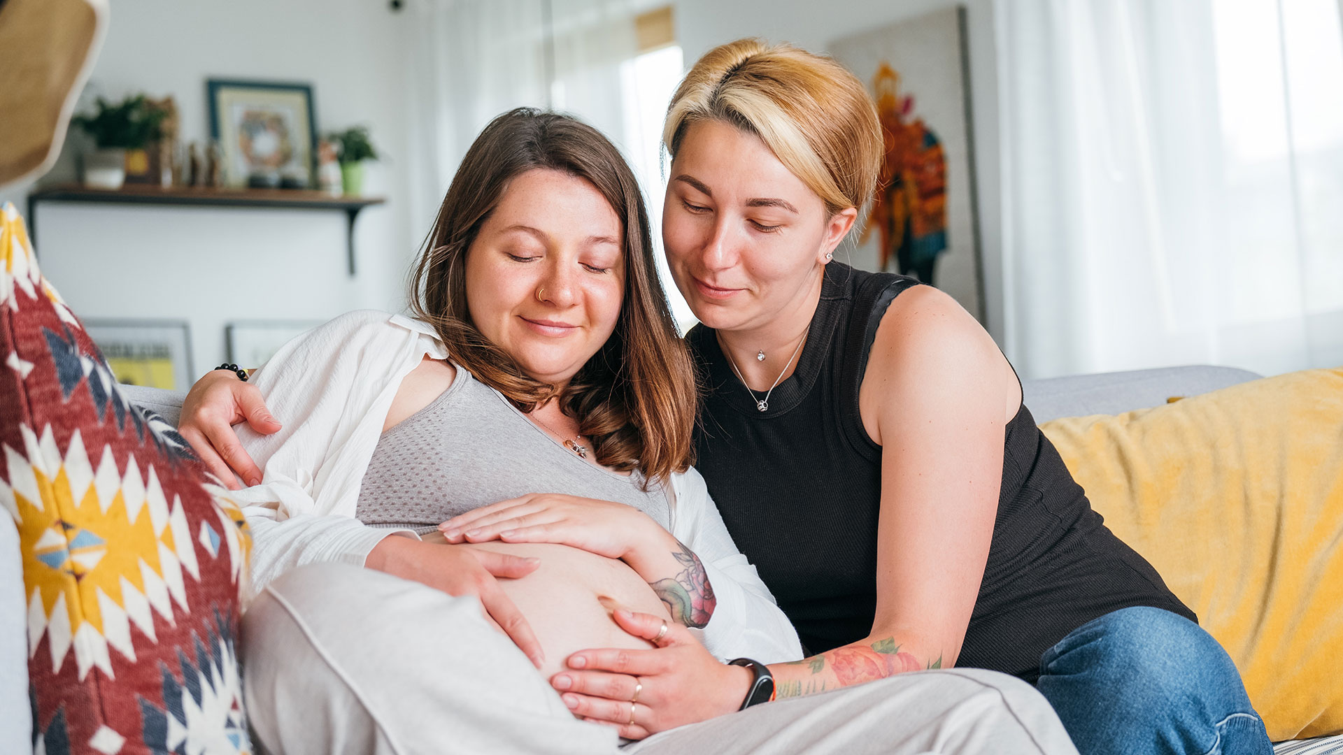 Zwei Frauen auf einer gelben Couch, die Frau links hat einen Babybauch, den beide mit den Händen berühren