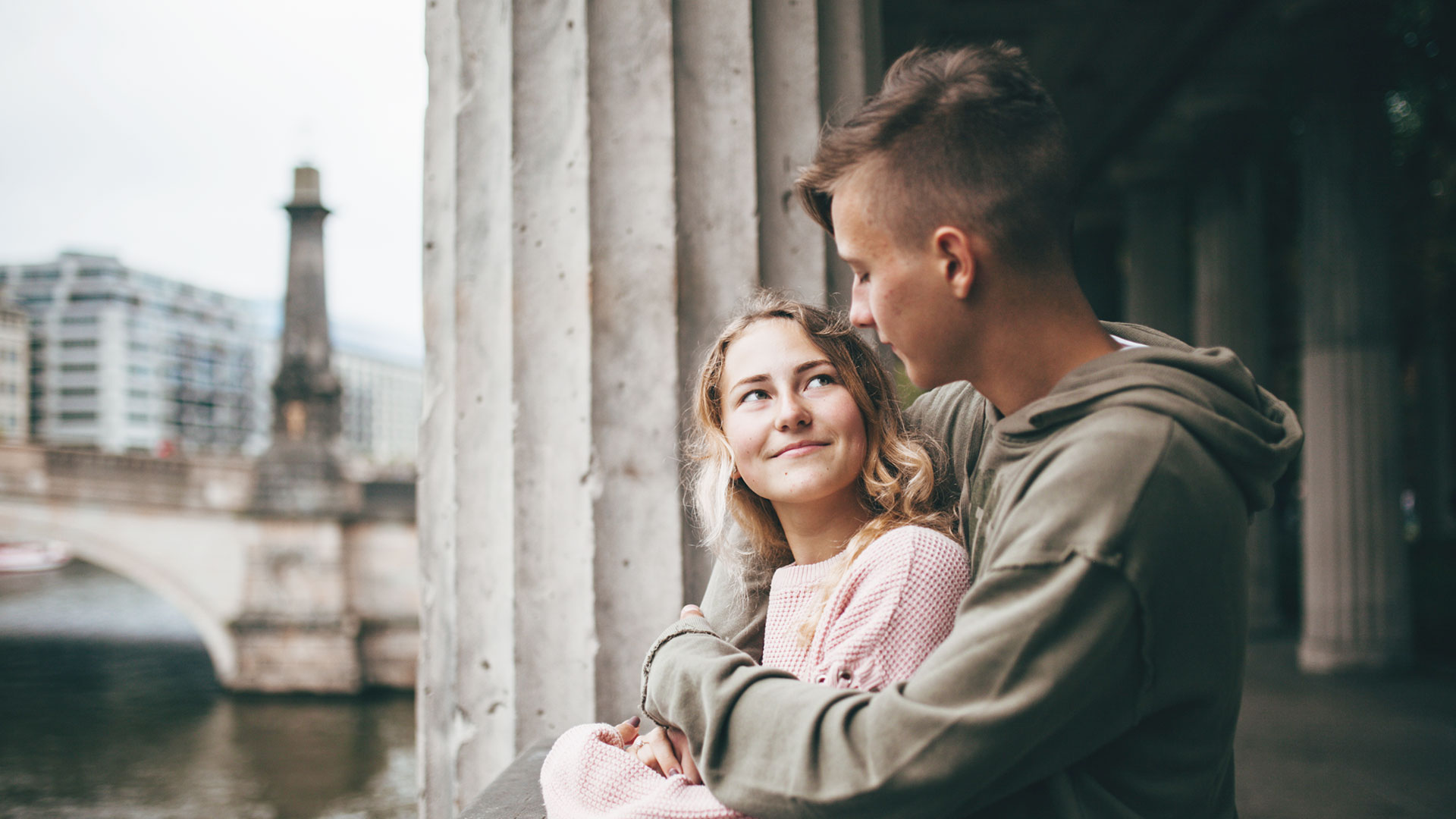 Junges Mädchen und Junge halten sich im Arm, dahinter eine Stadt