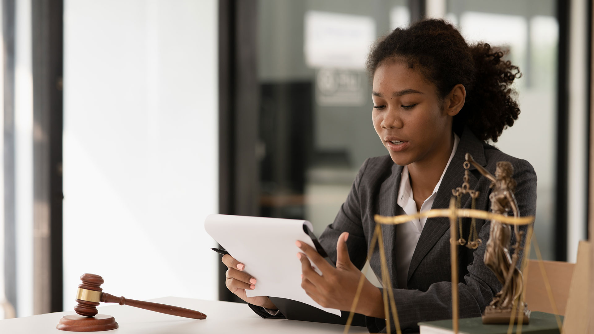 Junge Frau an einem Schreibtisch, blickt auf Papier ein ihrer Hand, vor ihr steht eine Justitia Waage