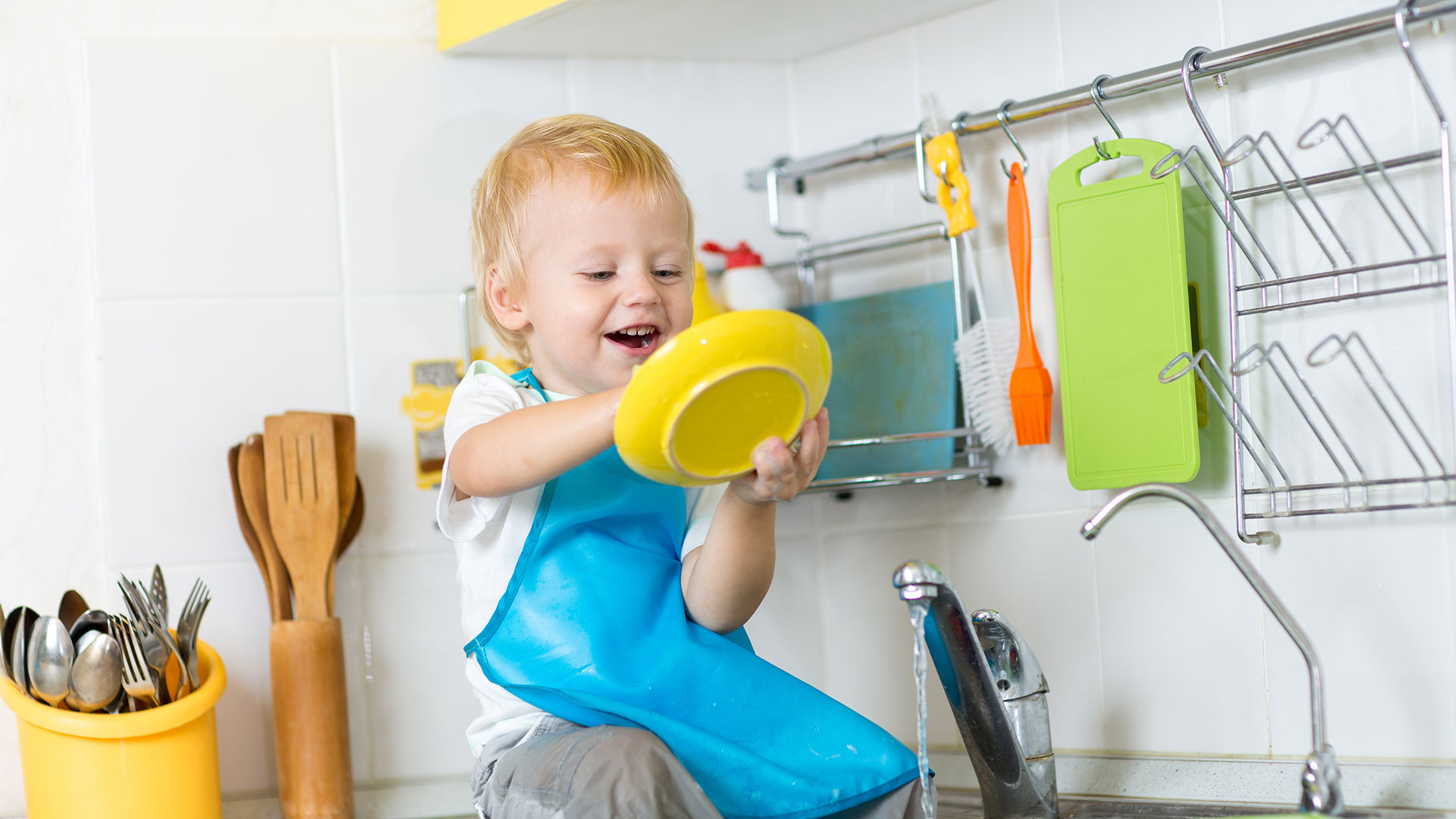 Blonder kleiner Jung sitzt am Rand eines Küchenbeckens und trocknet mit einem Geschirrtuch kindgerechte Plastikteller