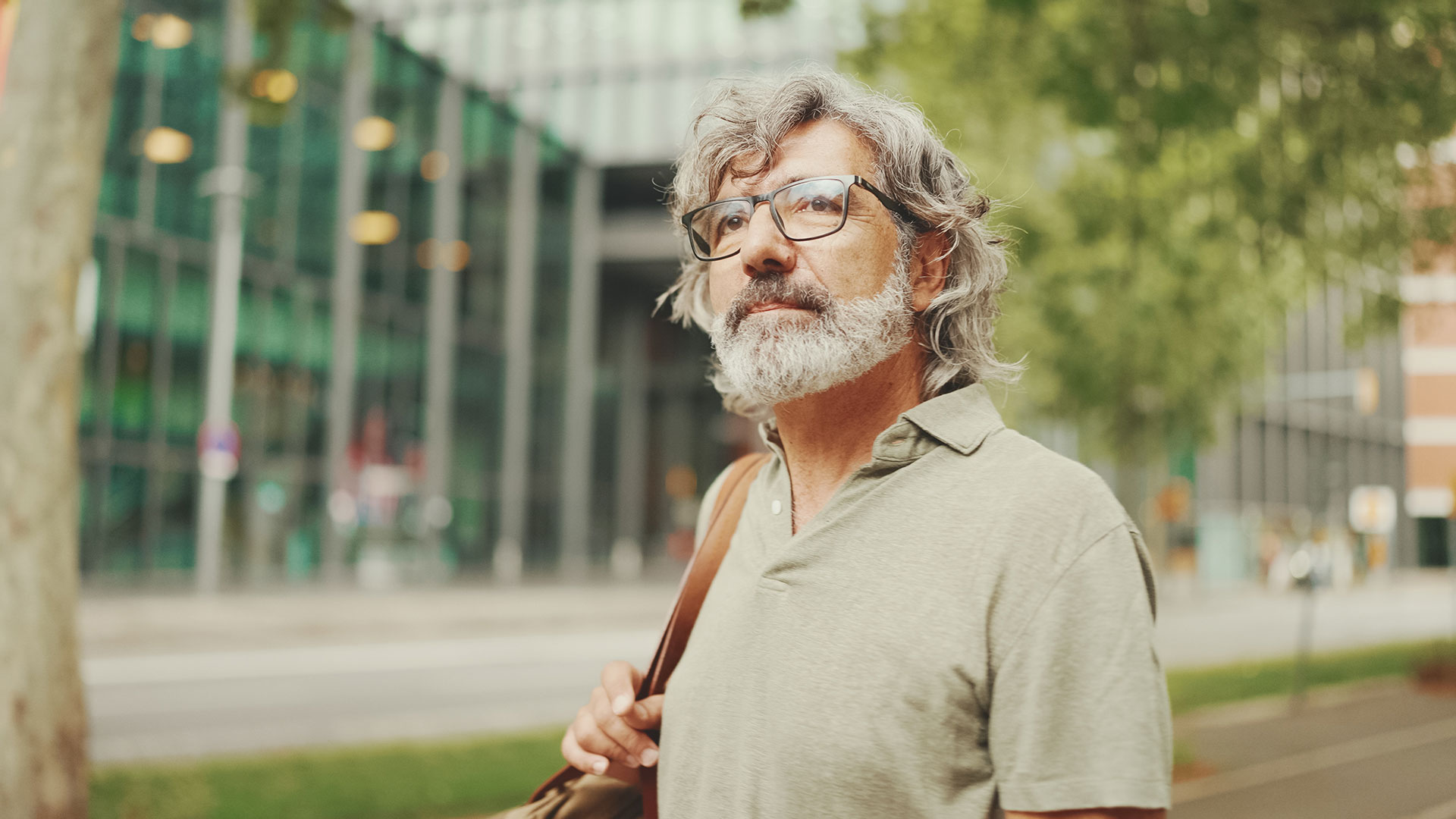 Mittelalter Mann mit Brille, grauem wallenden Haar und einem melierten Bart schaut nachdenklich, hinter ihm eine Glasfront eines städtischen Gebäudes