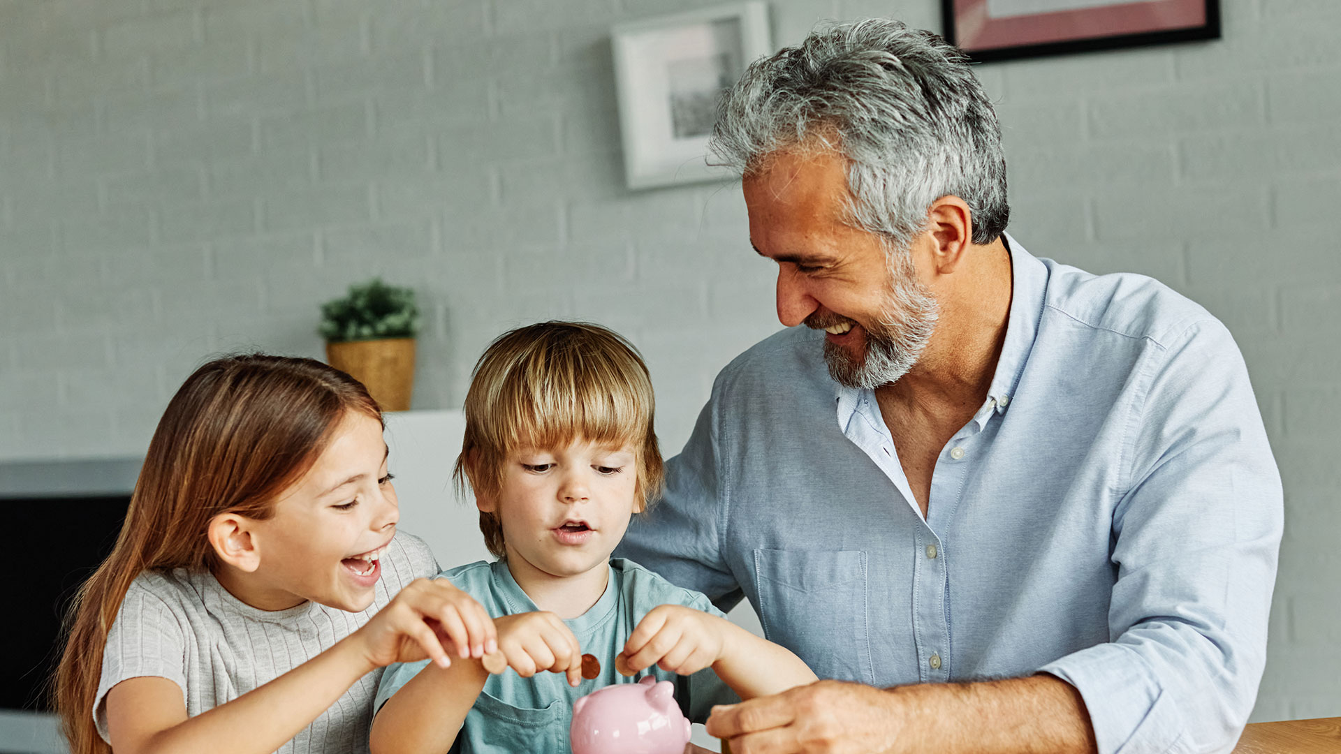 Ein Mann sitzt mit einem Jungen und einem Mädchen am Tisch, die Kinder befüllen freudestrahlend ein Sparschwein