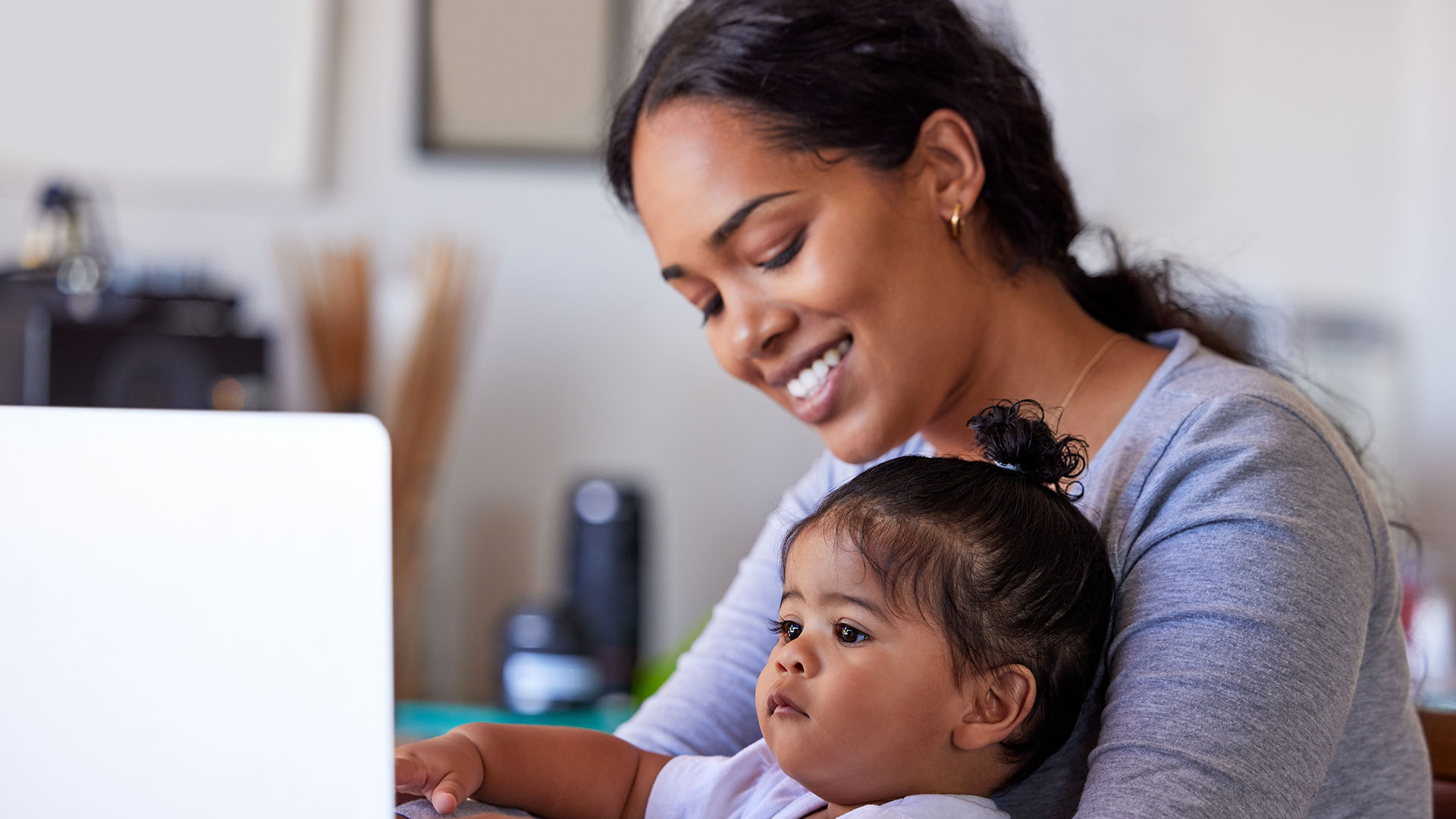 Junge Mutter arbeitet mit ihrem Baby auf dem Schoß am Laptop.