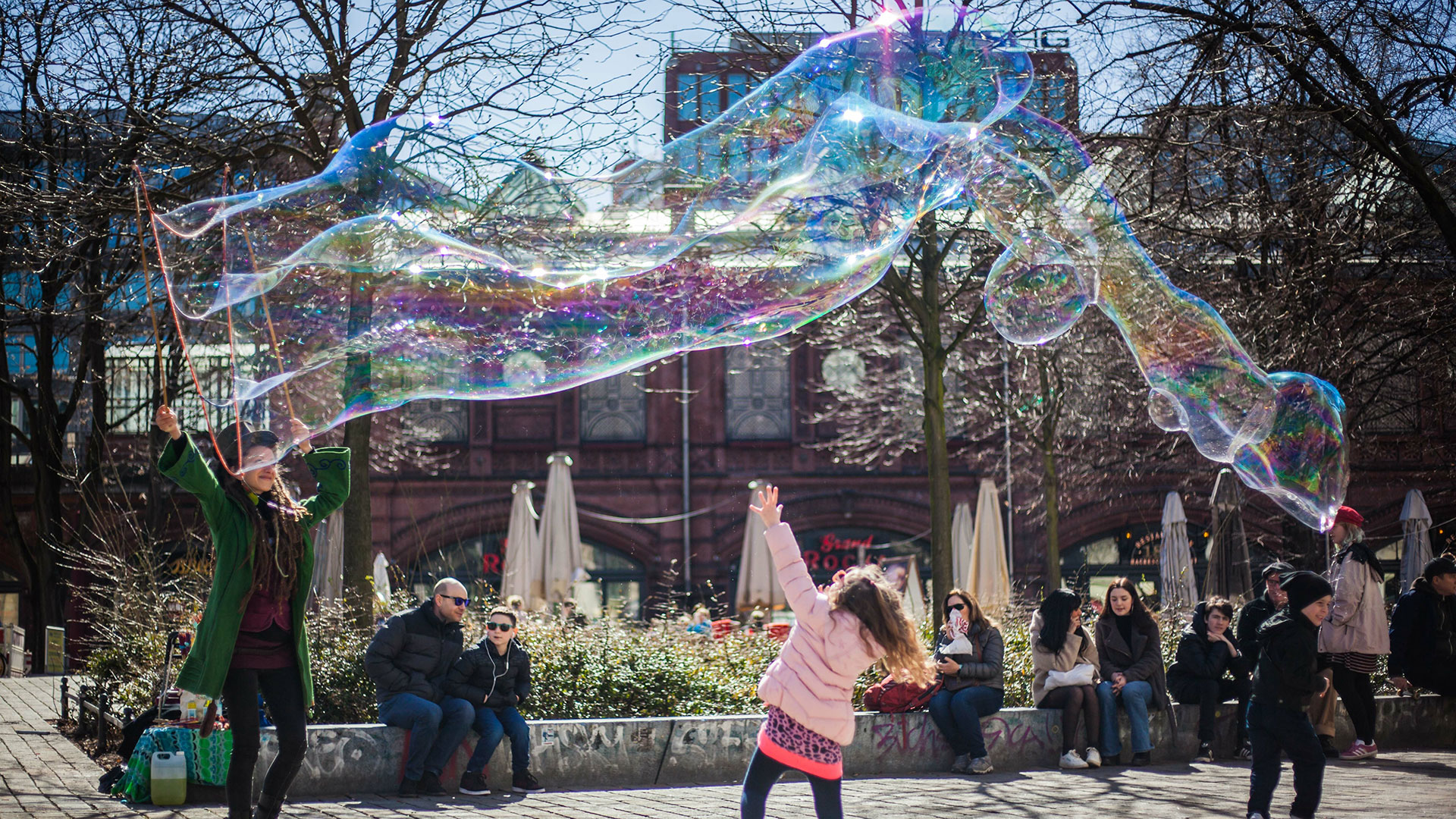 Menschen halten sich bei Sonnenschein auf einem städtischen Platz auf. Eine Frau macht riesige Seifenblasen, ein Mädchen tanzt darunter hindurch
