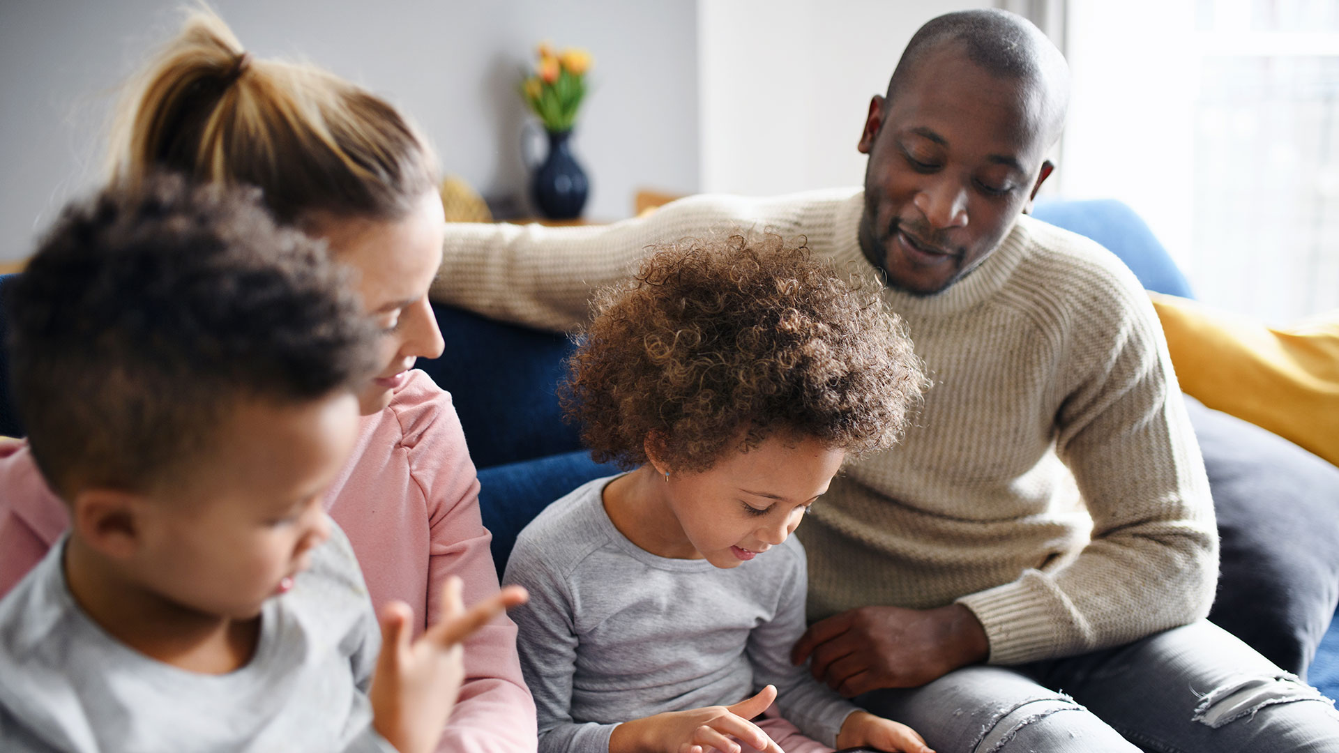 Eine multiethnische Familie sitzt gemeinsam auf dem Sofa. Ein Kind spielt mit einem Tablet-Computer.