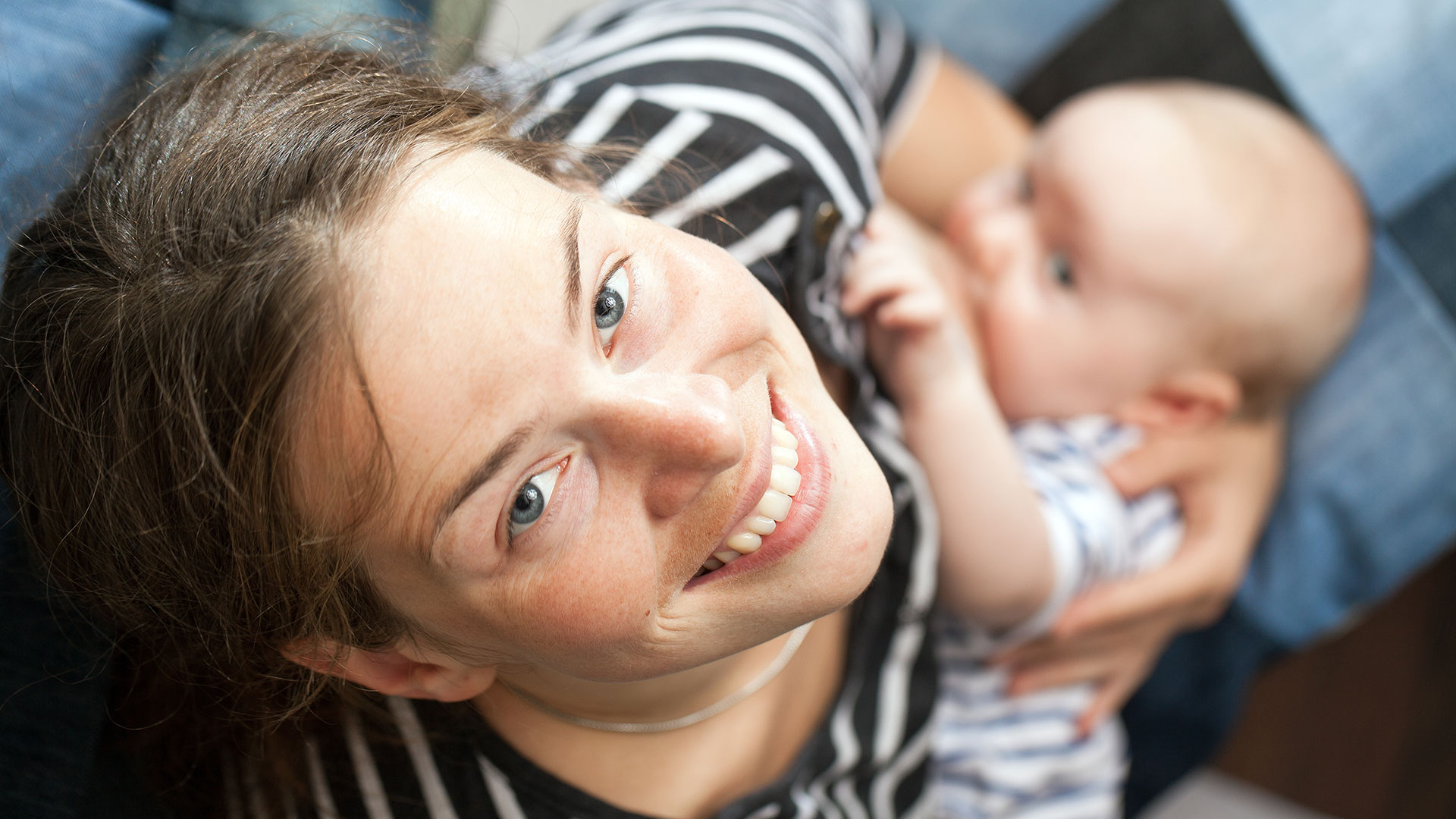 Eine junge Mutter stillt ihr Baby und blickt dabei glücklich in die Kamera.