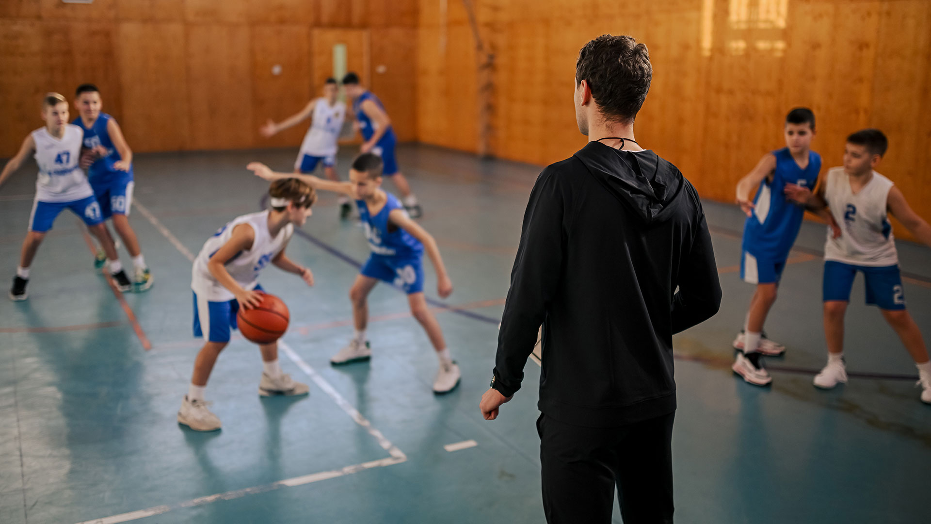 Eine Gruppe Jungs spielt Basketball in einer Sporthalle. Ihr Trainer steht am Spielfeldrand. 