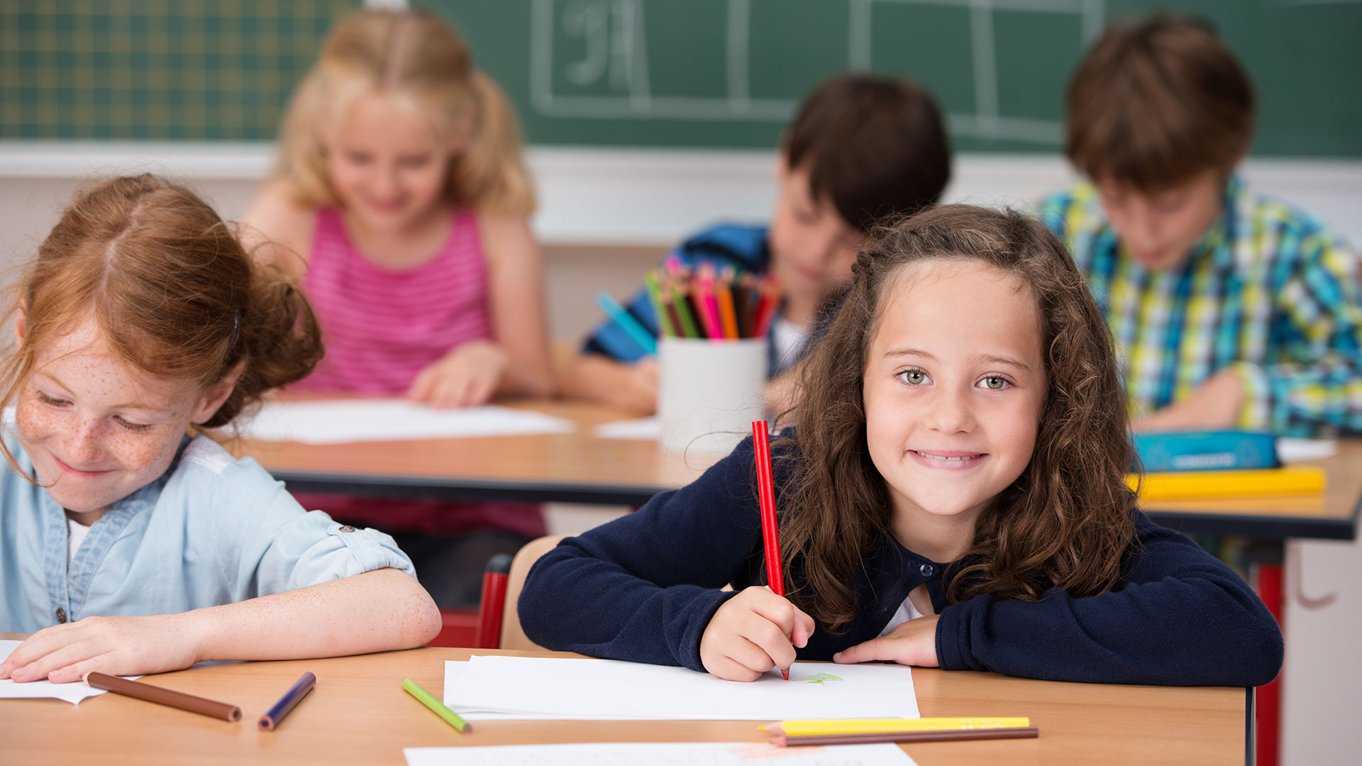 Kinder sitzen an Schreibtischen in einem Klassenzimmer und schreiben, ein Mädchen blickt auf und lächelt