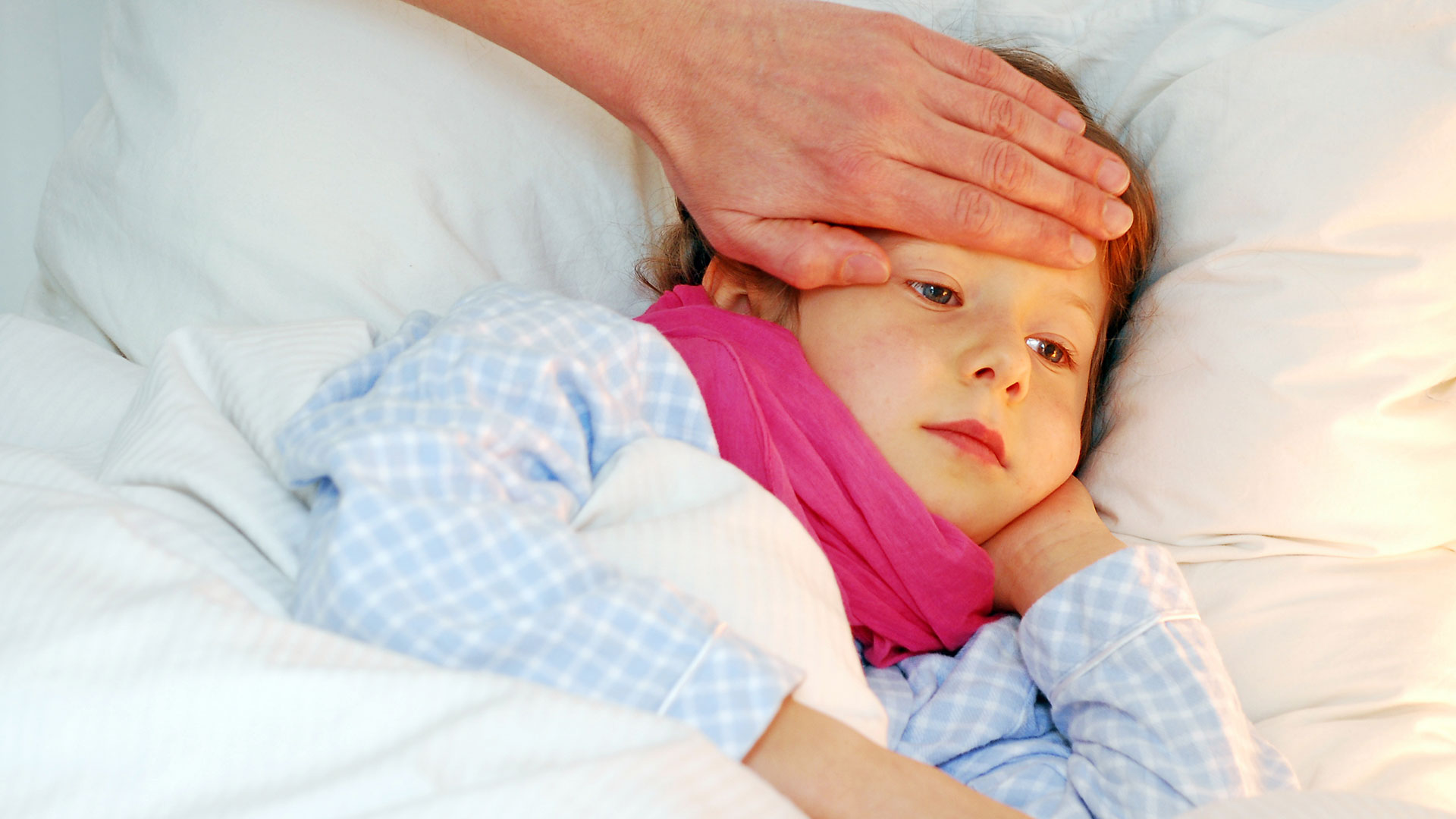 Ein Kind mit einem Schal liegt im Bett. Eine erwachsene Hand fühlt an der Stirn das Fieber.