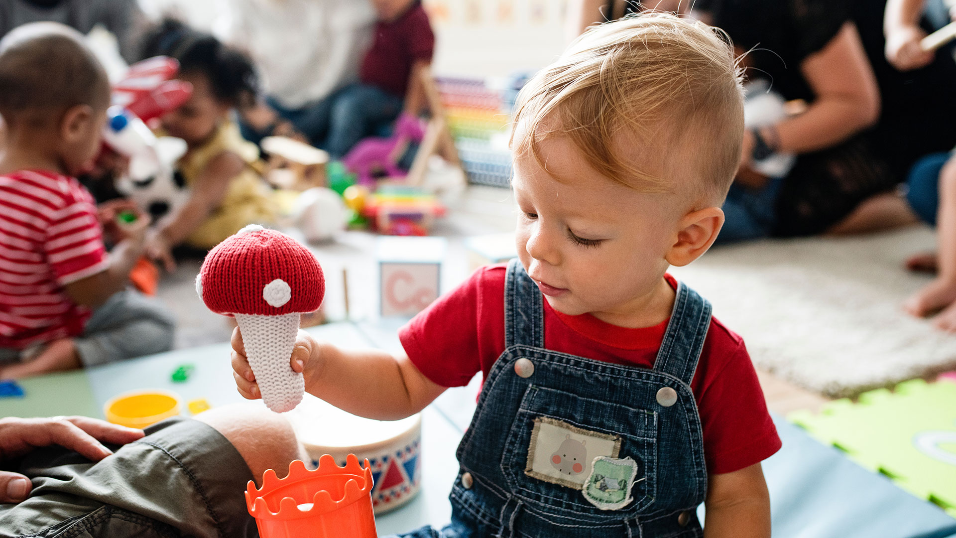 Ein Kleinkind in Latzhosen spielt in einer Kita mit einem gehäkeltem Pilz.