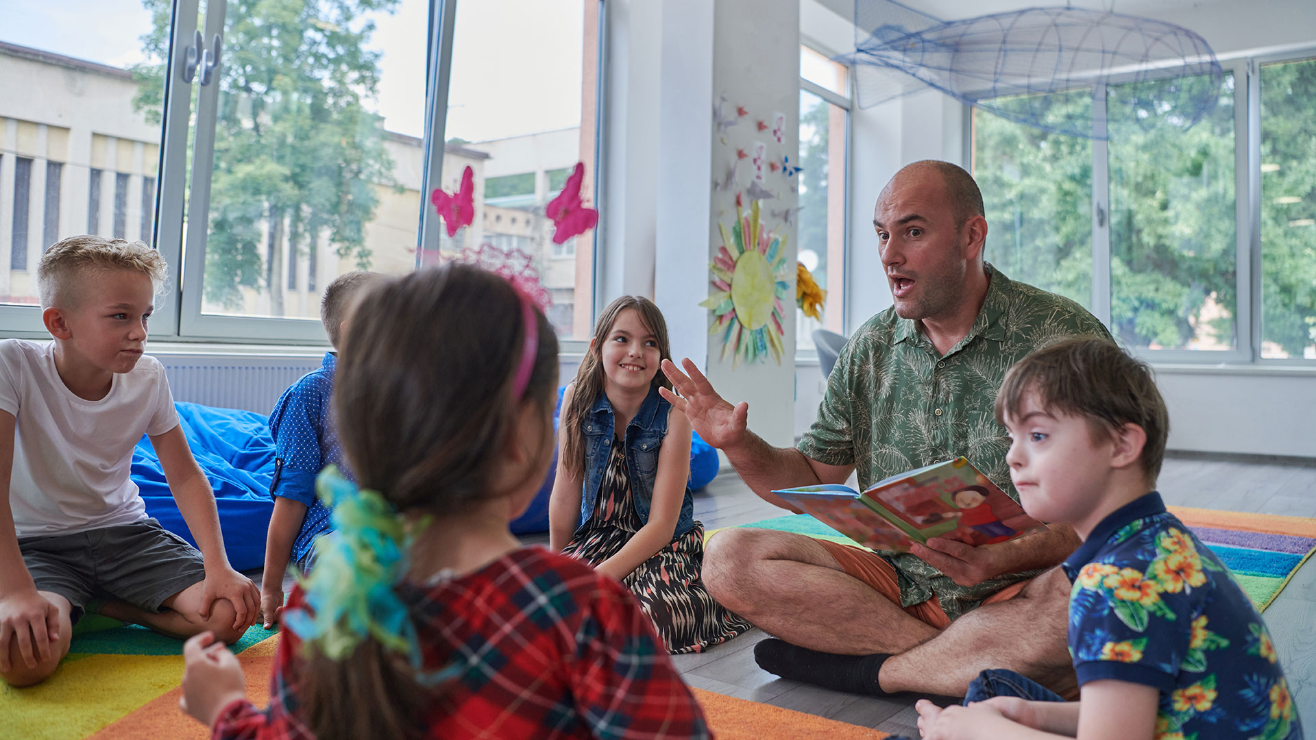 Ein Mann sitzt mit einer Gruppe Grundschulkinder auf dem Boden und liest vor.