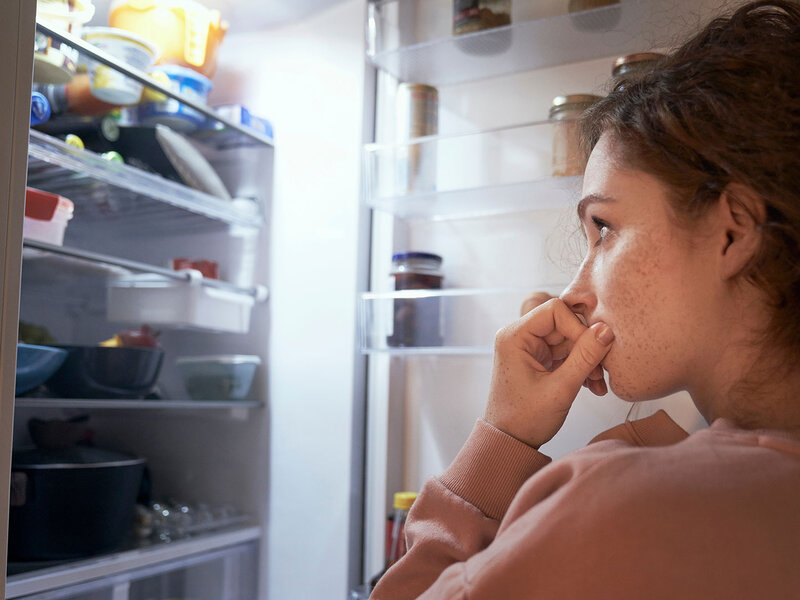 Junge Frau schaut unentschlossen in den geöffneten Kühlschrank.