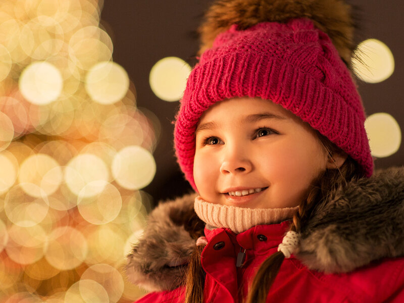Ein Mädchen mit roter Winterkleidung steht mit leuchtenden Augen auf einen abendlichen beleuchteten Weihnachtsmarkt.