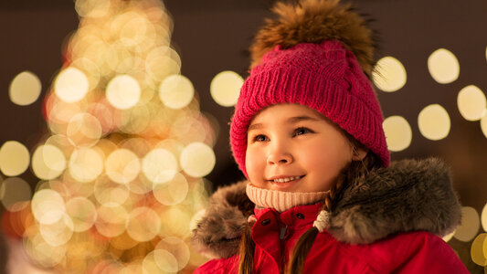 Ein Mädchen mit roter Winterkleidung steht mit leuchtenden Augen auf einen abendlichen beleuchteten Weihnachtsmarkt.