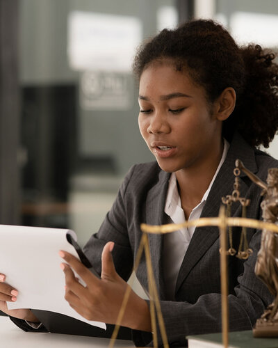 Junge Frau an einem Schreibtisch, blickt auf Papier ein ihrer Hand, vor ihr steht eine Justitia Waage