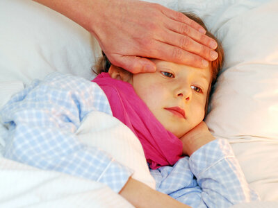 Ein Kind mit einem Schal liegt im Bett. Eine erwachsene Hand fühlt an der Stirn das Fieber.