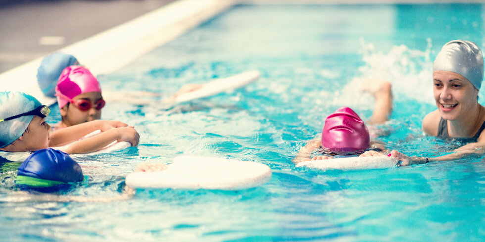 Kinder mit Badekappen und Schwimmbrettern bei einem Schwimmkurs in einem Hallenbad.