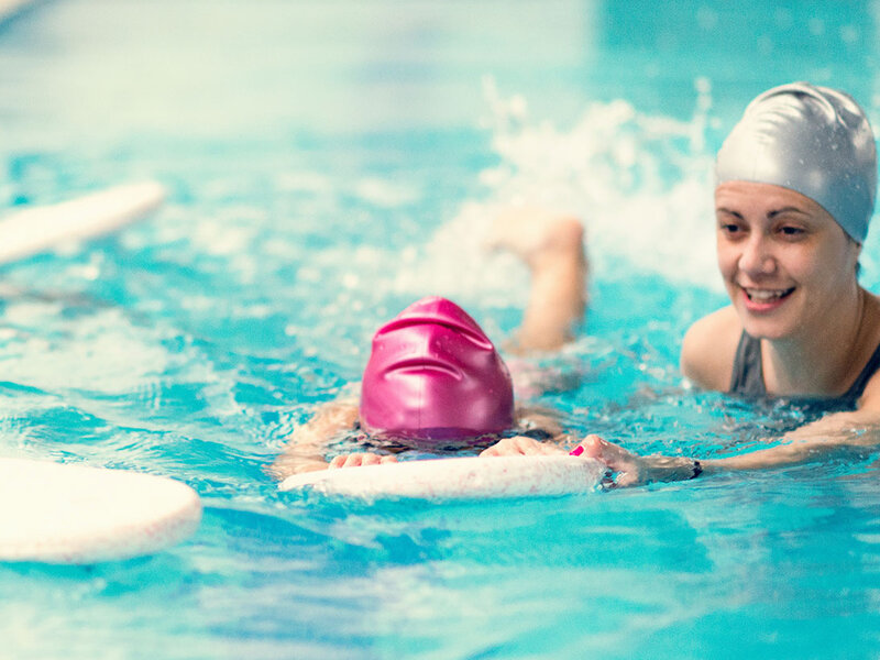 Kinder mit Badekappen und Schwimmbrettern bei einem Schwimmkurs in einem Hallenbad.