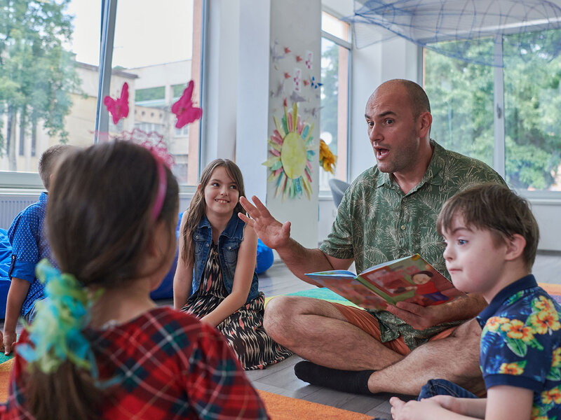 Ein Mann sitzt mit einer Gruppe Grundschulkinder auf dem Boden und liest vor.