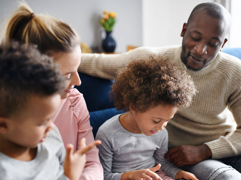 Eine multiethnische Familie sitzt gemeinsam auf dem Sofa. Ein Kind spielt mit einem Tablet-Computer.