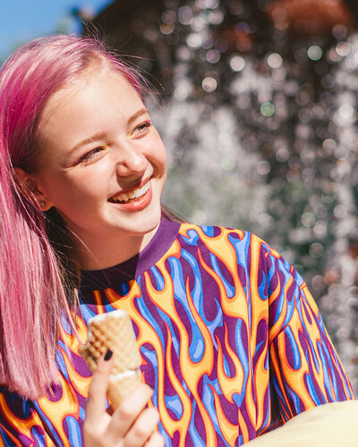 Mädchen mit pink gefärbtem Haar lächelt, hinter ihr ein Springbrunnen und blauer Himmel