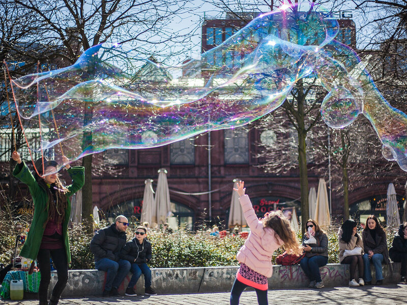 Menschen halten sich bei Sonnenschein auf einem städtischen Platz auf. Eine Frau macht riesige Seifenblasen, ein Mädchen tanzt darunter hindurch