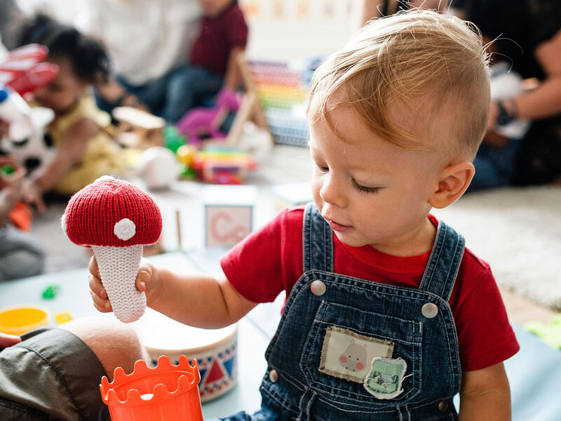 Ein Kleinkind in Latzhosen spielt in einer Kita mit einem gehäkeltem Pilz.