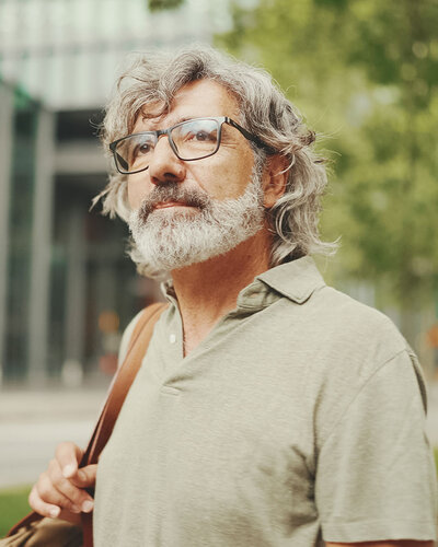 Mittelalter Mann mit Brille, grauem wallenden Haar und einem melierten Bart schaut nachdenklich, hinter ihm eine Glasfront eines städtischen Gebäudes