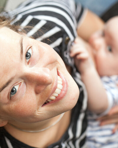 Eine junge Mutter stillt ihr Baby und blickt dabei glücklich in die Kamera.