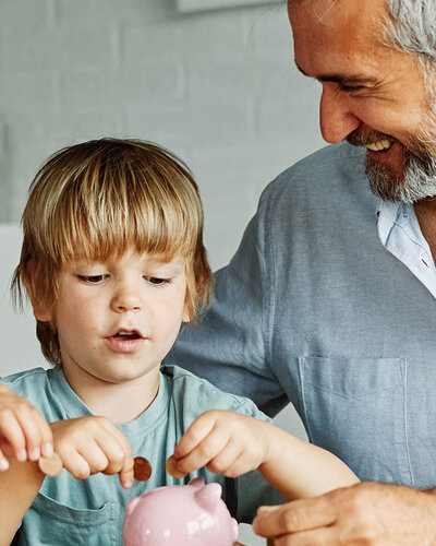 Ein Mann sitzt mit einem Jungen und einem Mädchen am Tisch, die Kinder befüllen freudestrahlend ein Sparschwein