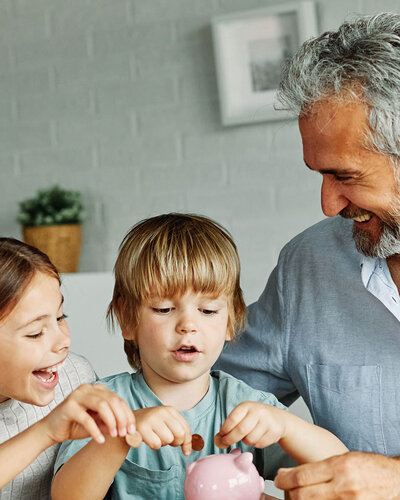 Ein Mann sitzt mit einem Jungen und einem Mädchen am Tisch, die Kinder befüllen freudestrahlend ein Sparschwein