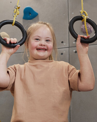 Ein lächelndes Mädchen mit Down Syndrom greift Ringe vor einer Kletterwand