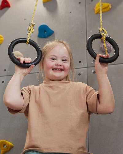 Ein lächelndes Mädchen mit Down Syndrom greift Ringe vor einer Kletterwand