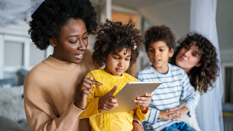 Zwei Frauen und ein Mädchen und ein Junge in deren Mitte schauen gespannt auf ein Tablet