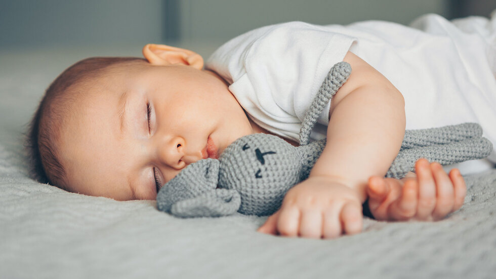 Ein schlafendes Baby hat einen grauen gehäkelten Hasen im Arm.