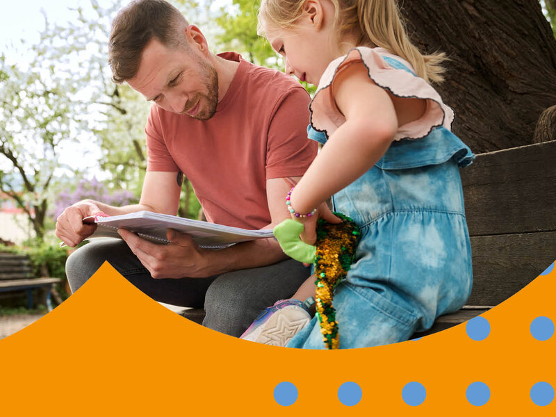 Ein Vater sitzt mit seiner Tochter auf einer Bank auf einem Spielplatz und kontrolliert die Hausaufgaben.