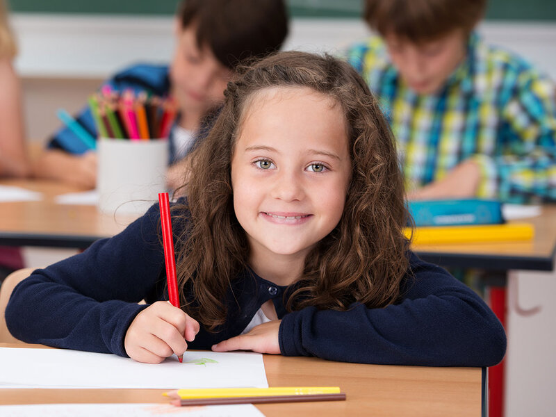 Kinder sitzen an Schreibtischen in einem Klassenzimmer und schreiben, ein Mädchen blickt auf und lächelt