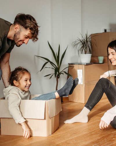 Ein junger Mann schiebt ein Kind, dass in einem Umzugskarton sitzt, durch eine leeren Wohnraum. Eine Frau sitzt daneben auf dem Parkettboden und schaut freudig zu