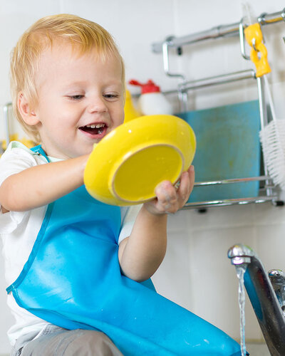 Blonder kleiner Jung sitzt am Rand eines Küchenbeckens und trocknet mit einem Geschirrtuch kindgerechte Plastikteller