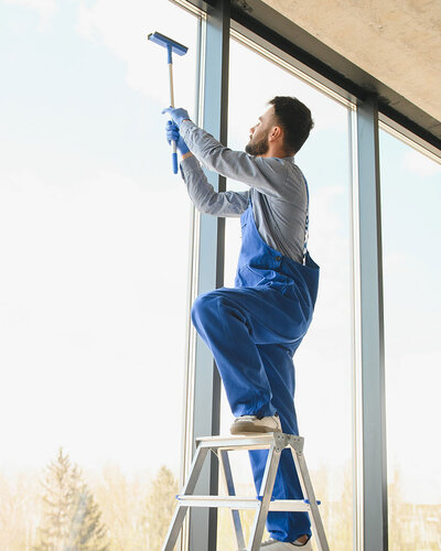 Mann an einer großen Fensterfront, steht auf einer Leiter und putzt ein Fenster