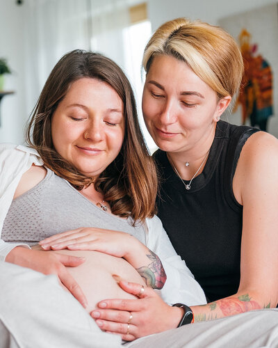 Zwei Frauen auf einer gelben Couch, die Frau links hat einen Babybauch, den beide mit den Händen berühren