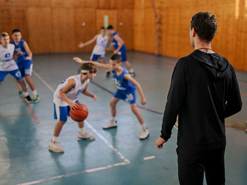 Eine Gruppe Jungs spielt Basketball in einer Sporthalle. Ihr Trainer steht am Spielfeldrand. 